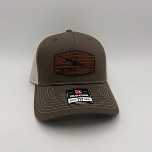 May include: Brown and beige trucker hat with a patch featuring an American flag design and the text "AC-130J". The hat has a curved brim and an adjustable strap. The brand "Richardson" is visible on a tag.