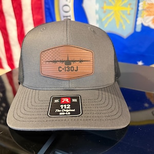 May include: Gray trucker hat with a black mesh back and a brown leather patch. The patch features an American flag design and the text "C-130J". The hat has a curved brim and a tag that reads "112 The Original MD-LG".