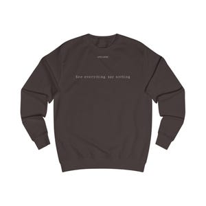Può includere: Una felpa marrone scuro con la scritta "APEX LOGIC" sopra la frase "See everything, say nothing" in un semplice font. La felpa ha maniche lunghe e girocollo.