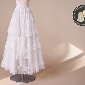 Op de afbeelding: Een witte, gelaagde kanten rok, tentoongesteld op een mannequin. De rok heeft een brede elastische tailleband en meerdere lagen delicate kant, waardoor een volle, vloeiende silhouet ontstaat. Een cirkelvormig logo met de woorden "Timeless Classic Wardrobe" is zichtbaar.