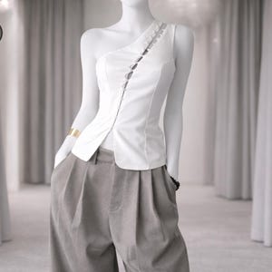 Könnte beinhalten: Ein weißes One-Shoulder-Top mit Knopfleiste und Zierleiste. Dazu eine graue Bundfaltenhose. Ein goldenes Armband und ein schwarzes Armband sind sichtbar. Das Bild ist vor einem neutralen Hintergrund aufgenommen.