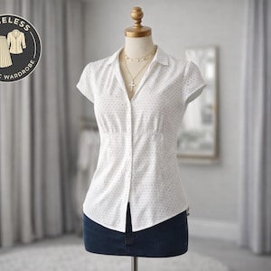 Puede incluir: Blusa blanca de manga corta con pequeños lunares oscuros, exhibida en un maniquí. La blusa tiene cuello y cierre de botones. Se ve un collar dorado con un colgante de cruz. El maniquí lleva una falda vaquera azul oscuro. La imagen incluye un logo "Timeless Classic Wardrobe".