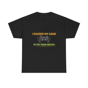 May include: Black t-shirt featuring the text "I paused my game to fix your server." A grey games controller is positioned below the text. The text is in yellow and green.