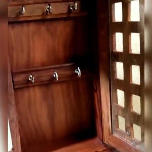 Pode incluir: Um armário de chaves de madeira com uma porta articulada, com um acabamento castanho-avermelhado rico. O interior tem duas filas de ganchos prateados para guardar chaves. A porta tem um design decorativo recortado.
