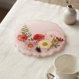 Puede incluir: Un posavasos redondo de resina de color rosa pálido con un borde ondulado y festoneado. El posavasos está incrustado con varias flores prensadas, incluyendo flores rojas, rosas y blancas, junto con hojas de helecho verdes en la superficie.