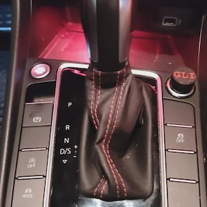 Może przedstawiać: Czarna dźwignia zmiany biegów w samochodzie z czerwonymi przeszyciami. Konsola posiada przyciski do parkowania, trybów jazdy i klimatyzacji. Przycisk oznaczony "GLI".