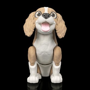 May include: A 3D printed figurine of a sitting dog with tan and white fur. The dog has large brown eyes, a black nose, and a pink tongue hanging out. It features floppy ears, a white chest and paws, and a textured surface throughout the body.