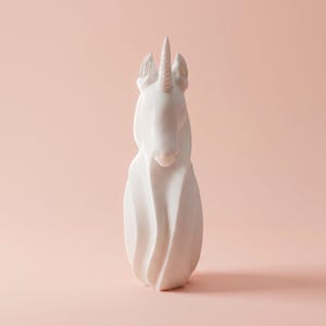 May include: A white unicorn sculpture with a spiraling body and a detailed horn. The sculpture is set against a soft pink background, highlighting its elegant form. This decorative piece is ideal for home decor.