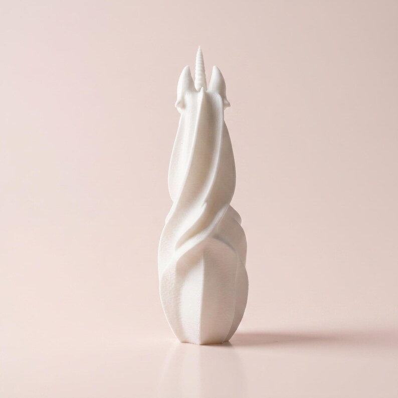 May include: A white, abstract unicorn sculpture with a spiraling body and a single horn. The sculpture has a smooth, matte finish and is set against a soft pink background. The design is modern and minimalist, suitable for home decor.