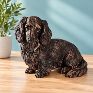 May include: A bronze-colored figurine of a long-haired dachshund dog. The sculpture features detailed fur texture with dark brown and copper highlights, depicting the dog in a seated position with its head turned slightly toward the front.