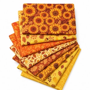 Peut inclure: Une pile de carrés de tissu pliés présentant des motifs sur le thème de l'automne. Les designs incluent des tournesols jaunes sur fond brun foncé, des feuilles d'érable orange, des glands bruns et divers motifs botaniques dorés et orangés.