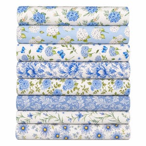 Könnte beinhalten: Ein Stapel aus acht Stoffballen mit verschiedenen blau-weißen Mustern. Die Designs umfassen blaue Hortensien auf Weiß, weiße Hortensien auf Hellblau, blaue Schmetterlinge und Blumen, florale Ornamente, Rosen, Ranken und Gänseblümchen.