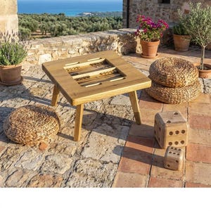 Puede incluir: Una mesa de backgammon de madera con una superficie de juego cuadrada y cuatro patas. Dos otomanas redondas tejidas y dos dados de madera están junto a la mesa. La escena está ambientada al aire libre en un patio de piedra con plantas y vistas al mar.
