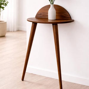 Puede incluir: Una mesa auxiliar de estilo moderno de mediados de siglo con respaldo de media luna y tres patas anguladas. La mesa es de madera oscura y sostiene un jarrón blanco con flores coloridas. La mesa mide 71 cm de alto.