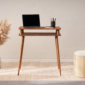 May include: A wooden desk in a mid-century modern style, with a laptop, pen holder, and notepad. The desk has a lower shelf and angled legs. A vase of pampas grass and a round stool with a light-coloured cushion are also in the image.