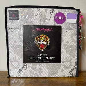 Op de afbeelding: Een 6-delige laken-set van Ed Hardy in een doorzichtige plastic opbergtas. De stof heeft een grijs en wit schakelpatroon met hart- en tijgerafbeeldingen. Een zwart label toont een tijgerkop, het Ed Hardy-logo en de tekst 6-Piece Full Sheet Set.