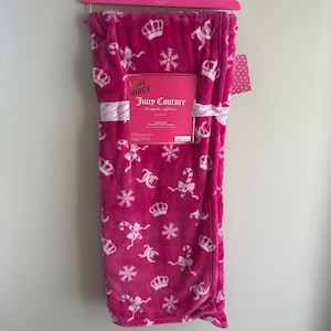 Op de afbeelding: Een fuchsia kleurige pluche deken met een herhalend patroon van witte kronen, sneeuwvlokken, zuurstokken en de letters "JC". De deken is voorzien van de labels "Juicy Couture" en "Love Juicy".