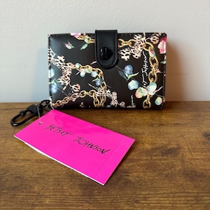 May include: Black wallet with a floral, chain, and leopard print design. The wallet has a black snap closure with a heart-shaped detail. A pink tag with the text "Betsey Johnson" is attached to the wallet.