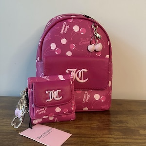 May include: A magenta backpack and wallet set with a cherry pattern and the text "Juicy Couture". The backpack has a front pocket and a cherry charm. The wallet has a snap closure. A pink tag with the brand name is also present.