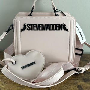 May include: A light pink handbag with a matching heart-shaped coin purse. The bag features black handles, a detachable shoulder strap, and the brand name "STEVE MADDEN" in black lettering. A tag with the brand name is attached.