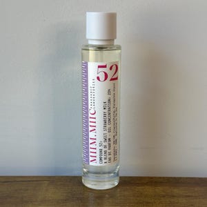 May include: A clear glass perfume bottle with a white cap. The label reads "52" and "Compound 52: A blend of sweet strawberry milk". The bottle contains a clear liquid. The label is purple and white.