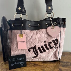 May include: A pink quilted Juicy Couture handbag with black detailing. The bag has a drawstring closure, gold-coloured hardware, and a heart-shaped charm. The brand name "Juicy" is embroidered in brown. A tag reads "Juicy Couture".
