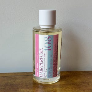 May include: A clear glass bottle of hair perfume with a white cap. The label features the text "HAIR PERFUME" and "108" in a pink and blue design. The bottle contains 3.4 oz of product.
