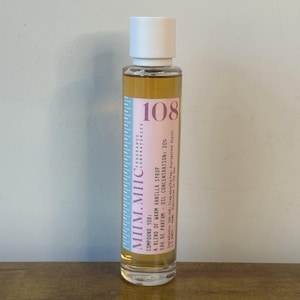 May include: A clear glass perfume bottle with a white cap. The label is pink and white with the number 108. The bottle contains a light amber liquid. Text on the label reads "Compound 108: A blend of warm vanilla syrup".