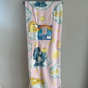 May include: Pink Care Bears twin blanket with Care Bears characters, clouds, and stars. The blanket is hanging on a black rod. The blanket has a tag that says "Twin Blanket".