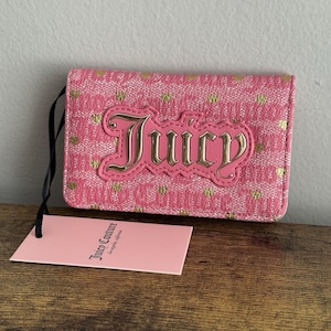 May include: Pink wallet with a gold heart pattern and the word "Juicy" in gold lettering. A pink tag with the words "Juicy Couture" is attached to the wallet with a black string. The wallet is on a wooden surface.