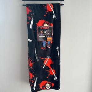 Può includere: Coperta in pile nera con un motivo di coltelli, schizzi di sangue rosso e il personaggio Chucky. La coperta ha un'etichetta con il logo Chucky e l'immagine del personaggio. La coperta è appesa a un'asta nera.