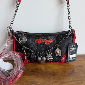 May include: A black Ed Hardy crossbody bag with a debossed skull pattern, red trim, and a silver chain strap. The bag features a red logo, multiple metal charms including a skull and heart, and a detachable red strap wrapped in plastic packaging.