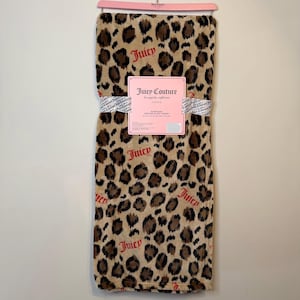 Op de afbeelding: Een bruin en beige deken met luipaardprint en het woord "Juicy" in rode letters. De deken is opgevouwen en bijeengehouden met een wit lint. Een roze label met "Juicy Couture" is bevestigd.