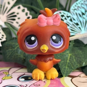 May include: A small, stylized brown bird figurine with a large head and oversized purple eyes. The bird features a bright yellow beak, a small pink bow on top of its head, and orange feet. The figure has a smooth, matte finish and a rounded body.