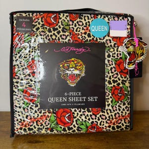 Peut inclure: Un ensemble de draps queen 6 pièces Ed Hardy dans un emballage en plastique transparent. Le tissu présente un motif léopard avec des roses rouges et des feuilles vertes. Une étiquette noire affiche le logo Ed Hardy, un tigre et le texte 6-PIECE QUEEN SHEET SET.