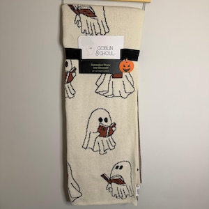 May include: A cream-coloured knit throw blanket featuring a repeating pattern of black-outlined ghosts reading brown books. A black paper band across the centre displays the text Goblin & Ghoul, Decorative Throw, and 127 cm by 152 cm.