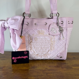 Pode incluir: Bolsa tote Juicy Couture rosa claro com um padrão acolchoado. A bolsa apresenta um fecho com cordão, ferragens prateadas e um laço decorativo. Um berloque em forma de coração e um berloque de chave pendem da alça. A bolsa tem texto bordado e um design de coroa.