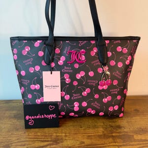 May include: Black tote bag with a pink cherry pattern and black handles. The bag features the text "Juicy Couture" and a pink logo. A pink tag and a small black card with text are also visible.