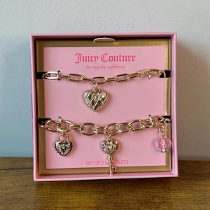 Könnte beinhalten: Ein Set aus zwei goldfarbenen Juicy Couture Armbändern. Eines der Armbänder hat einen Herzanhänger, das andere einen Schloss-, Schlüssel- und rosa Perlenanhänger. Die Armbänder werden in einer rosa Schachtel mit dem Juicy Couture Logo präsentiert.