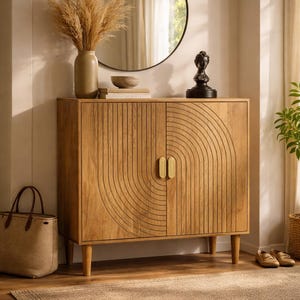 May include: A wooden cabinet with a carved, arched design on the doors, featuring brass handles. A round mirror hangs above it, with a vase of dried plants and a small sculpture on top. A woven tote bag and a rug are in the foreground.