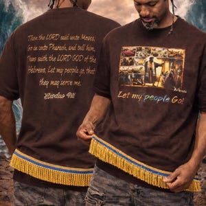 Könnte beinhalten: Braune T-Shirts mit religiösem Text und Bildern. Die Vorderseite des Shirts zeigt eine Grafik von Moses und die Worte "Let my people Go!". Auf der Rückseite des Shirts befindet sich ein Zitat aus Exodus. Der Saum des Shirts hat eine gelbe und blaue Fransen.