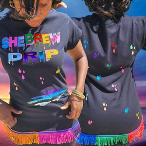 May include: Black t-shirts with the text "SHEBREW DRIP" in colorful, dripping letters. The shirts have a rainbow fringe at the bottom and colorful droplet designs. The image shows the front and back of the shirt.
