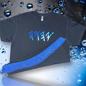 Puede incluir: Una camiseta gris oscuro con texto azul. El texto parece estar en un idioma extranjero. Una tira azul texturizada está drapeada en la parte inferior de la camiseta. El fondo es un degradado de azul y blanco con gotas de agua.