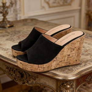 May include: Black peep-toe wedge sandals with cork-style platforms. The upper is made of black suede-like material. The wedge heel is approximately 4 inches high. The sandals are displayed on a decorative table.