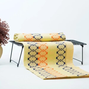 May include: A light yellow fabric runner featuring a repeating geometric pattern of dark blue and orange linked circles. The textile is folded and draped over a black wire rack, showcasing a consistent woven texture and a vibrant color palette.