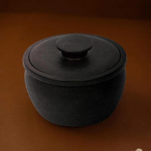 May include: A matte black, round stone bowl with a matching lid featuring a central circular handle. The bowl has a smooth, curved exterior and a thick rim, presenting a solid, dark appearance with a consistent, uniform finish throughout the piece.