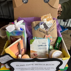 May include: A gift box labeled "Blind Date With A Book" filled with various items. Includes a book, a pen, a candle, a face mask, and other small gifts. The box is decorated with purple tissue paper and a white bow.
