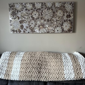 May include: A chunky knit blanket in beige and white stripes rests on a dark gray sofa. Above, a decorative wall art piece features a mosaic of silver and brown floral designs. The blanket is a cozy home decor item.