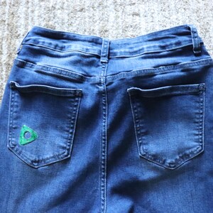 May include: Close-up of the back of blue denim jeans. The jeans have two back pockets and a small green triangle design on the left pocket. The jeans are on a light-coloured rug.