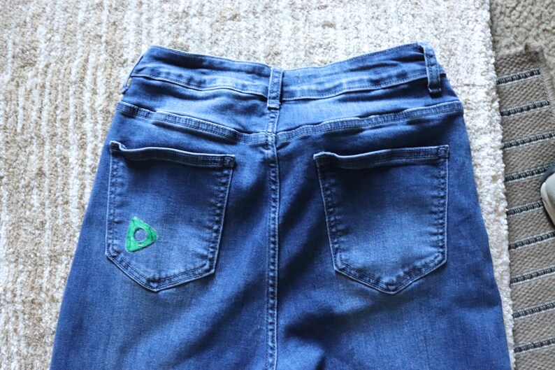May include: Close-up of the back of blue denim jeans. The jeans have two back pockets and a small green triangle design on the left pocket. The jeans are on a light-coloured rug.